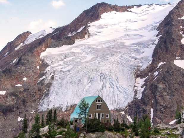 The Hut with its mint green arched roof, multiple windows and doorway framed in white sits surrounded by evergreen trees. A large glacier cascades down a steep slope behind the Hut.