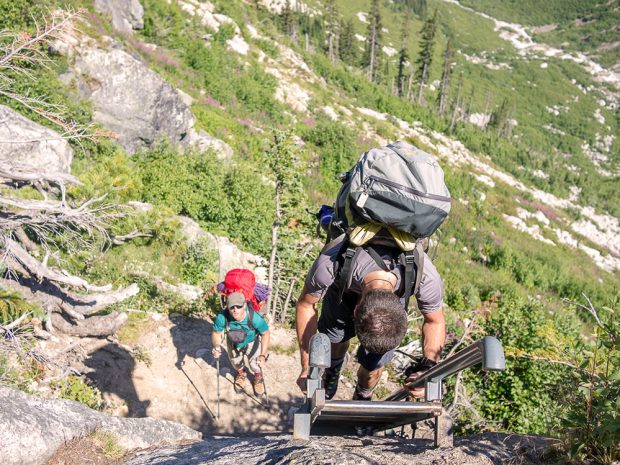 The photo taken from above looks down on a man climbing up the ladder with a grey hiking pack. Another man looks on from below wearing a green ball cap and a red backpack while holding on to his hiking poles.