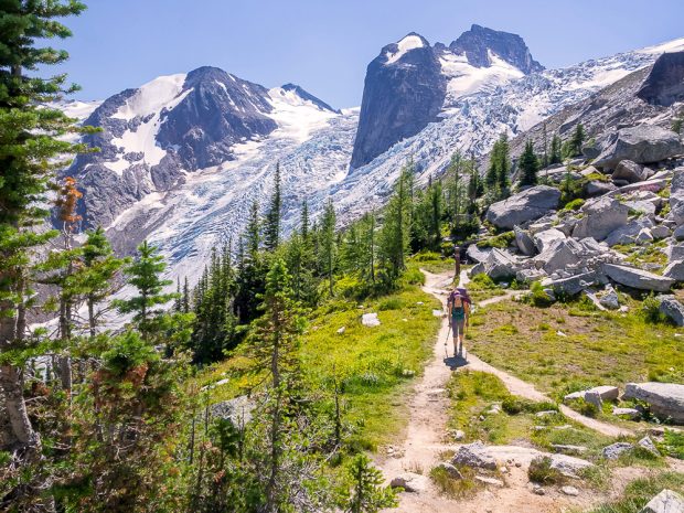 Glaciers surround jagged spires in the background. A women walks on the snowfree trail in the summer sunshine heading towards the glaciers and jagged peaks. Photo is taken from behind the woman walking.