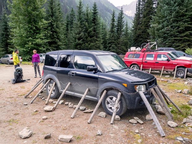 The blue vehicle is parked and the chickenwire mesh covers the bottom half of the car along with rocks and sticks. Two people stand near the parked vehicle chatting. Other cars can be seen with a similar setup.