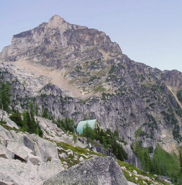 Looking up to the Conrad Kain Hut surrounded by evergreen trees and the large arete spire of the Bugaboos looms to the right of the hut.