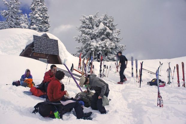 Three men sit on top of their backpacks to stay out of the deep snow are eating a snack in front of the hut. Skis and ski poles are stuck in the snow behind the group eating and another member walks towards the Hut entrance.