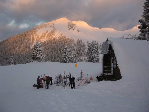 A group of skiers stand near ski equipment stuck in the snow near the front entrance of the Hut. The Hut is covered in a layer of snow and the sun is breaking through the dark grey clouds in the distance.