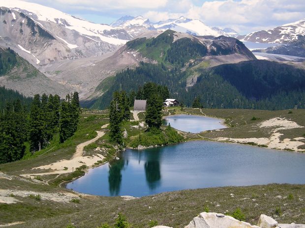 Two lakes sit in a green alpine valley and a gothic arch hut can be seen on the left of the smaller of the two lakes. Opal Cone can be seen in the distance and is free of snow.