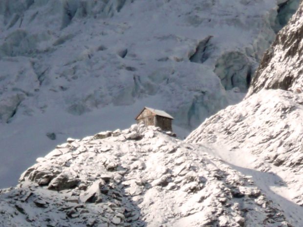 A stone shelter with an a-frame room sits on the top of a snow covered ridge and is bathed in sunlight. A large snow covered slope looms behind the shelter in the background.