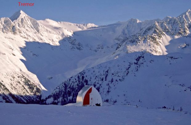 One half of the Himmelsbach Hut is bare of snow showing the red end-wall and silver aluminium siding covering the roof. The sun is shining on the snow- covered peaks in the background.