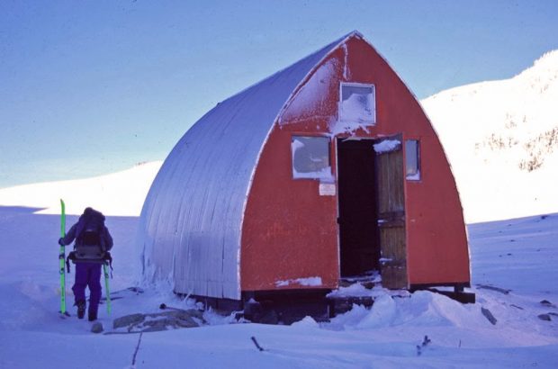 A man holding neon green touring skis is seen around the side of the hut and the front door is left ajar and the sun shines on the steep slopes behind.