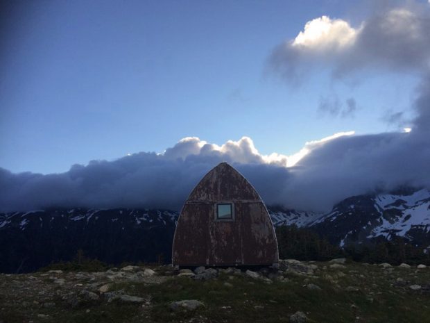 The Gothic arch Hut sits amongst a grassy meadow and loose boulders. Grey storm clouds cover the peaks in the distance.