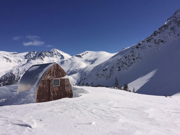 The end-wall of the hut shows severe signs of weathering and the snow and surrounding area is bathed in sunlight.