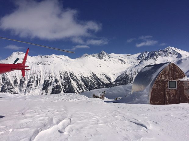 The tail and blade of the helicopter rotor can be seen at the left edge of the image and on the right edge of the photograph sits the weathered Himmelsbach Hut under a near cloudless blue sky.