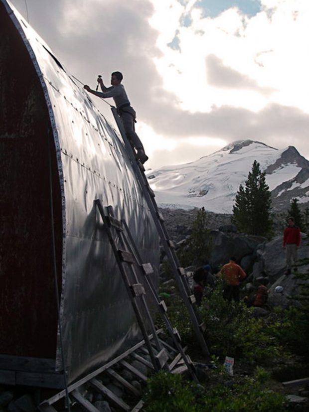 A man standing at the top of a hand built wooden ladder is inspecting the aluminum siding covering the roof. Other Club members can be seen standing behind near green evergreen trees. A steep snow covered slope looms in the background.