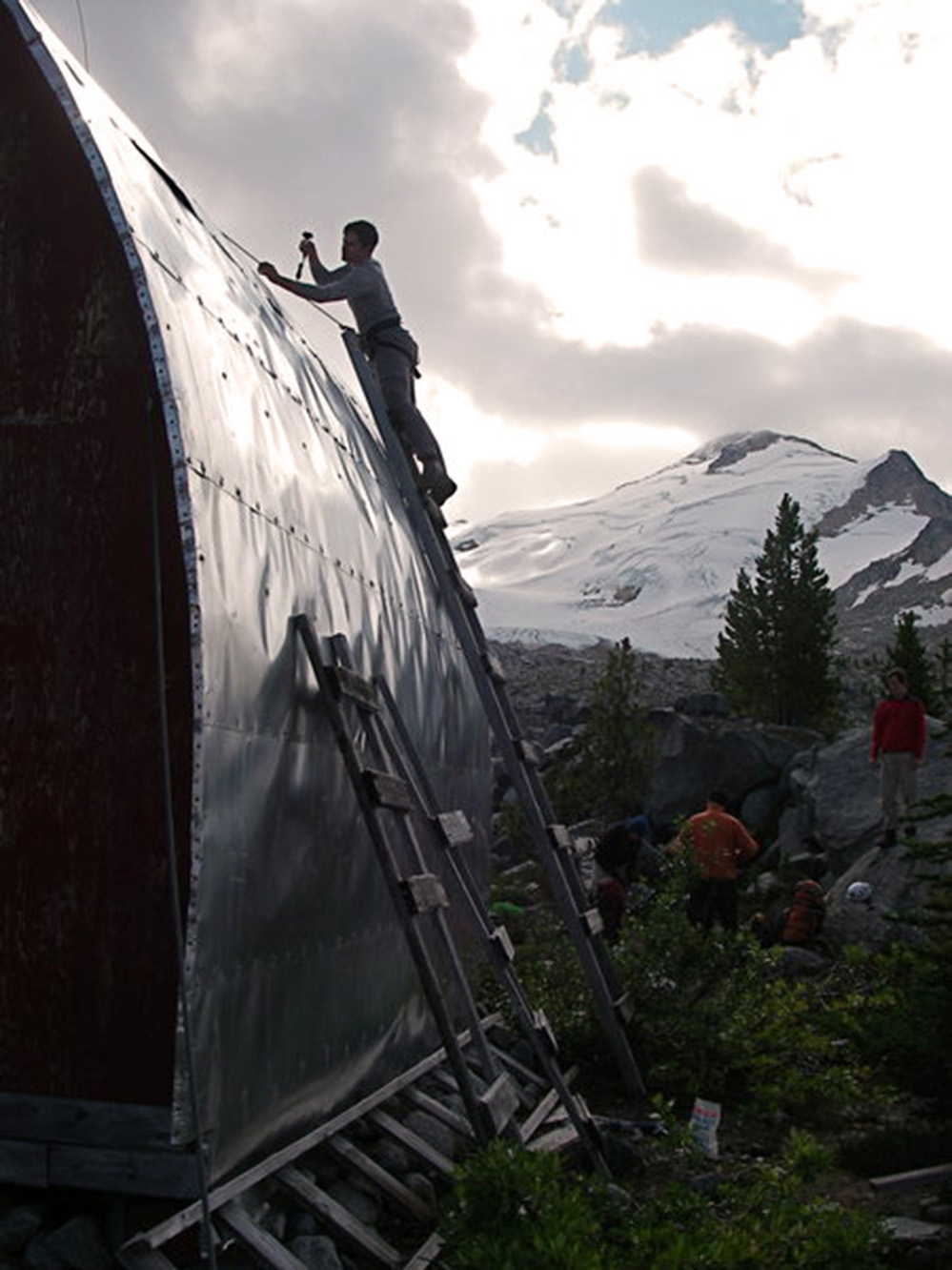 A man standing at the top of a hand built wooden ladder is inspecting the aluminum siding covering the roof. Other Club members can be seen standing behind near green evergreen trees. A steep snow covered slope looms in the background.