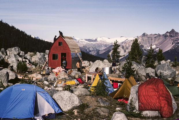 Tents and camping gear are in the boulder filled meadow in front of the Gothic Arch Hut. UBC-VOC members are busy working on completing the roofing and other aspects of the Hut construction. Snowy peaks are visible in the background.