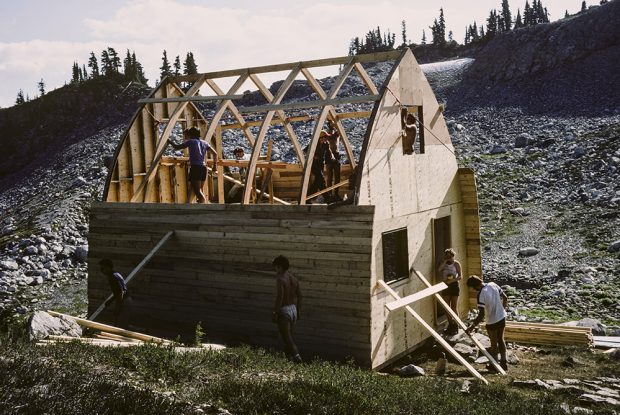 The Hut frame has been built and the tongue and groove siding is being added to both sides. Five members can be seen working on the second level and others are working on other tasks around the outside of the Hut.