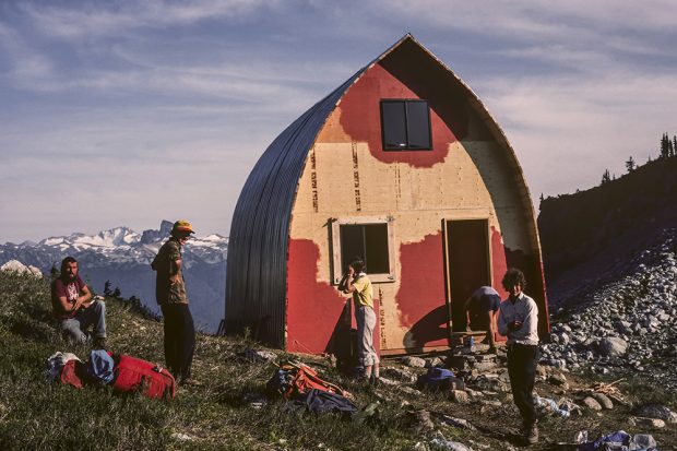 The Gothic Arch Hut framing is complete and painting has begun on the front end wall. Members of the work crew are out front of the hut standing in the sunshine.