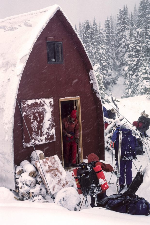 A skier is exiting the main entrance to the Hut and two other skiers are out front putting on their ski equipment. Snow is falling and the maroon coloured end wall of the hut stands out against the white snow.