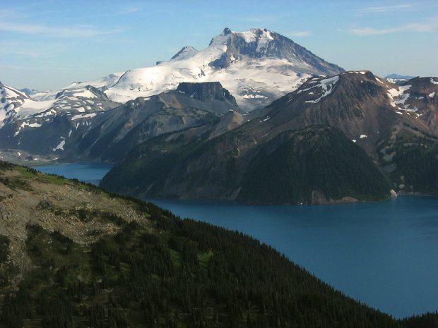 Wind ripples the deep blue colour of Garibaldi Lake and the glaciers above glisten in the sunshine. The brown and grey rock of the mountain peak in the background stands out in contrast to the deep green colour of the evergreen coloured slopes surrounding the lake.