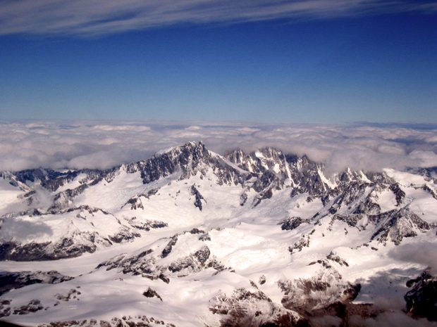 Blue sky extends in the distance above a larger glacier field leading to the base of Mount Waddington and the rock peak stands clear in the sunshine; other peaks surrounding Mount Waddington are blanketed by a bank of low lying grey storm clouds.