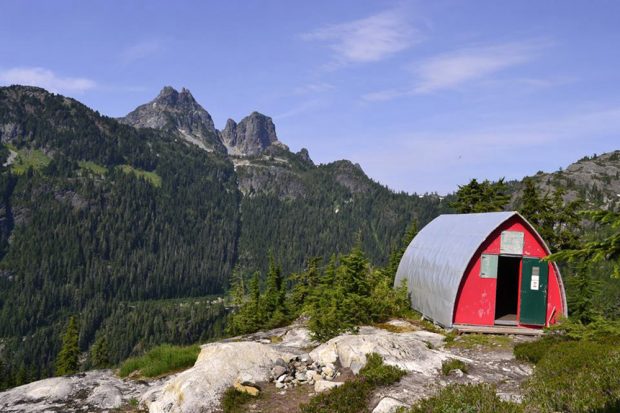 The photo was taken from an angle and the hut is located on the right hand-side with the front door open wide. The towering peaks across the valley from the hut stand out against the evergreen lined slopes.