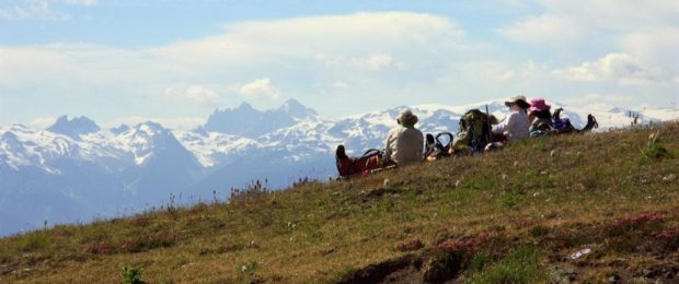 Three hikers sit near their backpacks all wearing sunhats looking out across at the distant mountain peaks. The peaks in the distance are covered in snow and clouds hang in the sky above them.