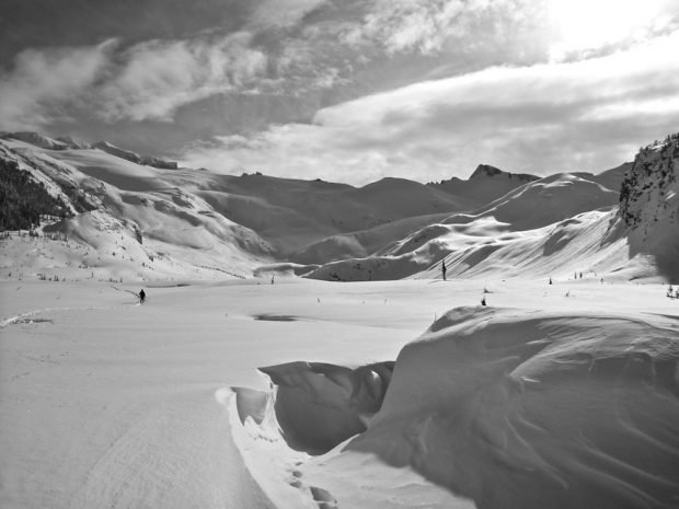 A black and white photograph of an alpine valley covered in snow with snow covered peaks in the background. A slight depression indicates where the Hut should be and a lone ski tourer is seen in the distance on the left.