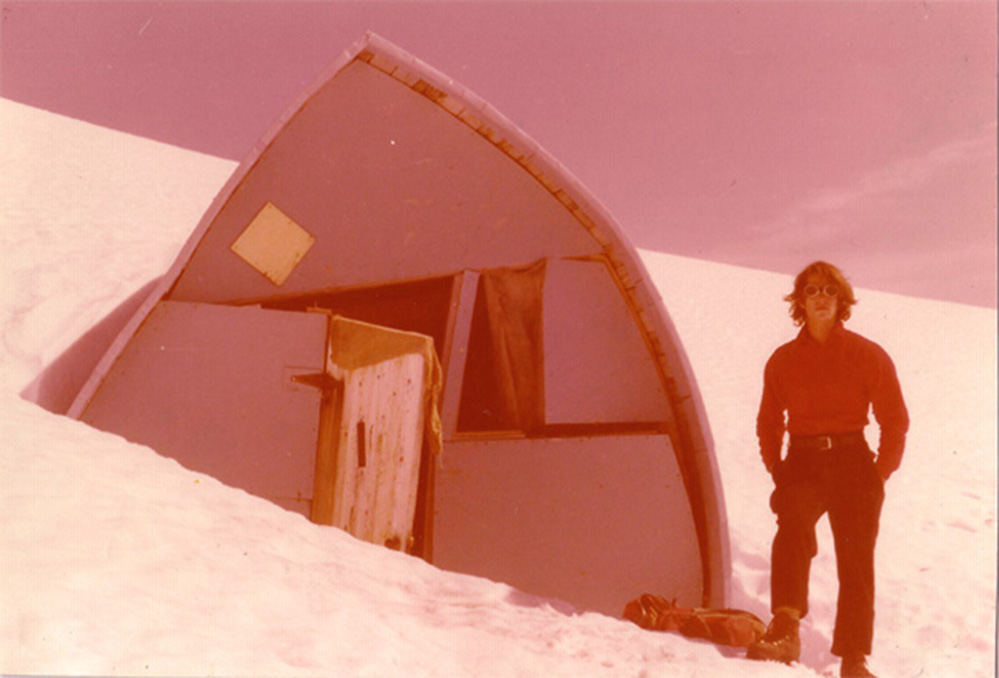 The pink-tinged photograph shows a Hut crushed by the weight of the snow and a man wearing red long-sleeve shirt and dark pants and brown hiking boots posing next to the Hut.
