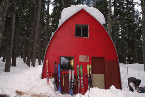The Hut entrance has been cleared of snow but a large tuft of snow still sits on top of the roof. Skis and ski poles lean up against the front end wall. A man hunched over wearing a white hat worked on clearing the entryway of snow.