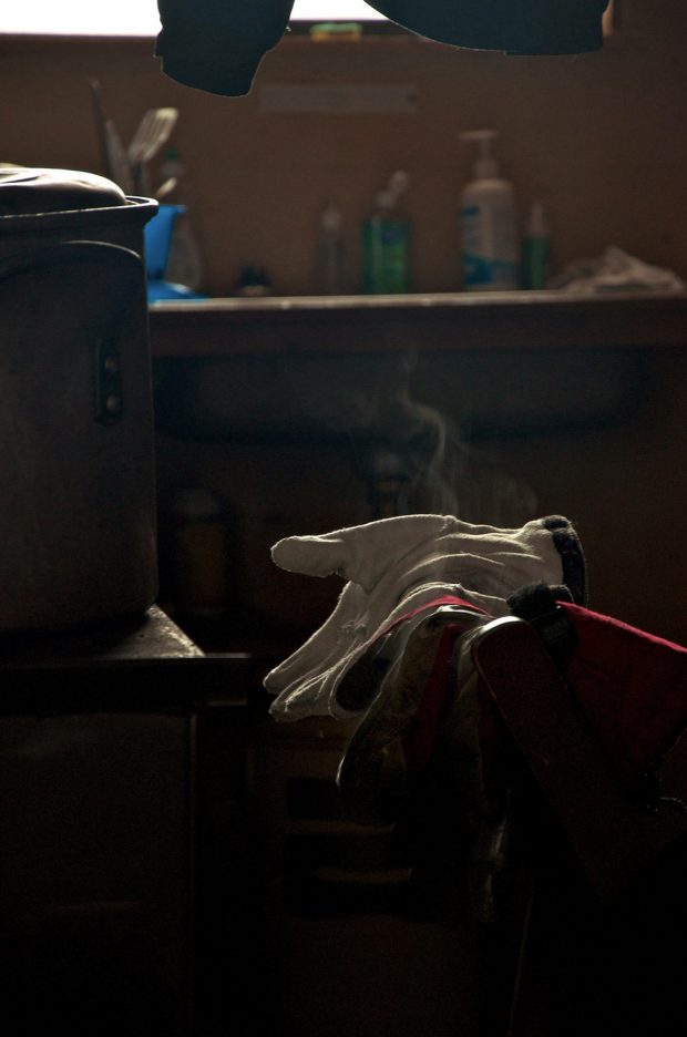 Cooking pot sits on the counter next to gloves sitting on top of clothes on a chair. Steam appears to be rising from the clothing and behind in the background, the sink lined with cleaning products and washed utensils drying in a blue plastic container.
