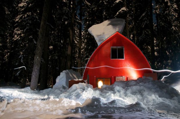 The Cabin surrounded by snow at night with the interior lights shining through the main floor window. A small clump of snow still rests on the arched roof.