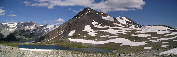 A small blue lake sits surrounded by a small ring of green grass and a large grey volcanic peak stands above it dotted with snow patches.