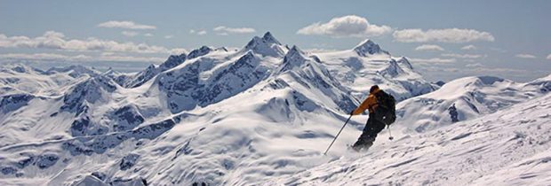 A skier in an orange jacket and black backpack is turning way from the photographer on a steep snowy slope with snow covered peaks in the distance.