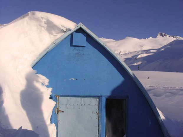 Blue front-wall of gothic arched hut nearly buried in snow, heaped up on one side and clear on the other. High snowy slopes and mountain peaks visible in background.