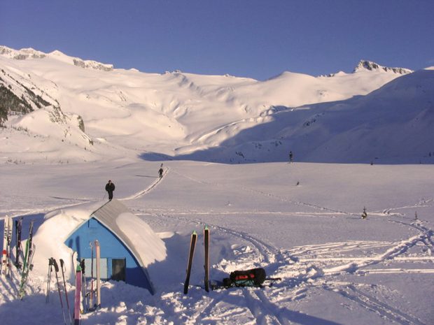 The Hut has been partially cleared of snow and skis and ski poles are stuck in the snow out front. Skiers can be seen in the distance making their way towards the Hut and the sun shines on the steep snowy slopes in the background.