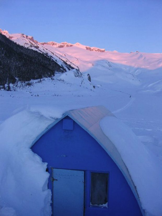 The blue end wall, front door and side window is free of snow and the sun is setting and leaving a pink and purple hue on the snowy slopes in the distance.