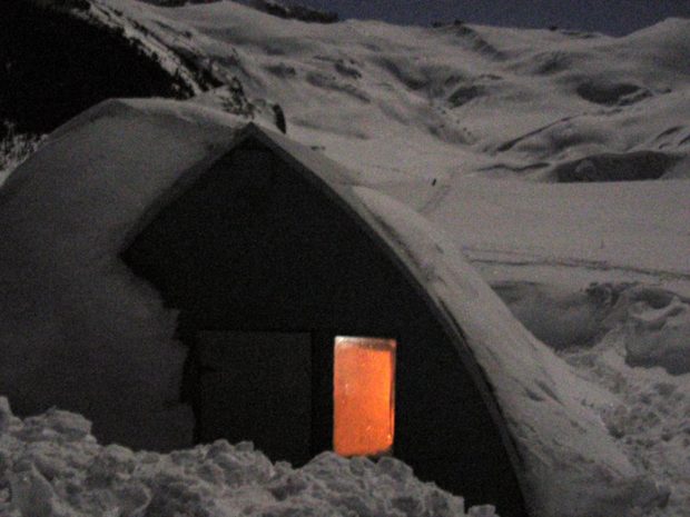 A hut partially buried in snow at night with the interior light reflecting in the uncovered window.