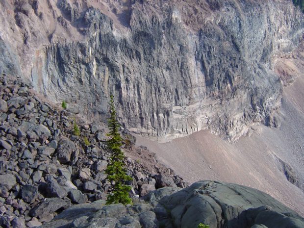 The bare red and grey rock wall illustrates the impact of glacial erosion. Below the wall are piles of sandy eroded rock and a small boulder field with a few small evergreen trees.