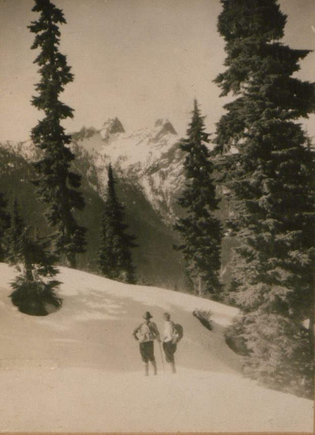 A couple stands on the snow slopes of the North Shore Mountains next to tall evergreen trees looking away from the camera to the two snowy peaks behind named the Lions.