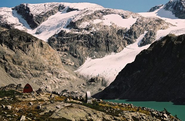 Wedge Mountain and its glacier loom large over the small Gothic arch Hut sitting in the rocky meadow below. The outhouse for the Hut is protected with aluminum siding and sits away from the Hut and near the Wedgemount Lake.