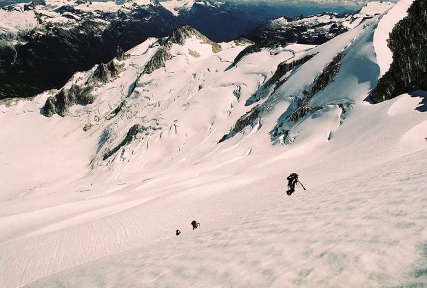 The photo looks down a large glaciated slope and the lead climber wearing a white helmet and a black backpack is partway up the slope with his left arm touching the snowy ground. Two other climbers farther down the slope are following the climber.