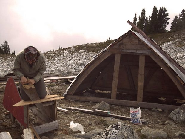 The remaining piece of the arched roof of the Brew Hut II sits on the ground. In the foreground, a man is busy hand-sawing a piece of wood.