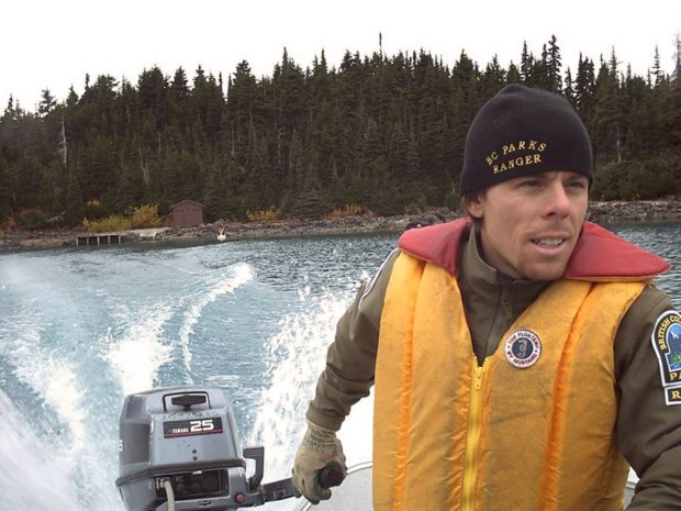 Portrait of a man wearing a yellow and red life jacket, while holding onto the handle of an outboard motor. The wake of the boat churns the water behind and off in the distance is the tree-lined shore.