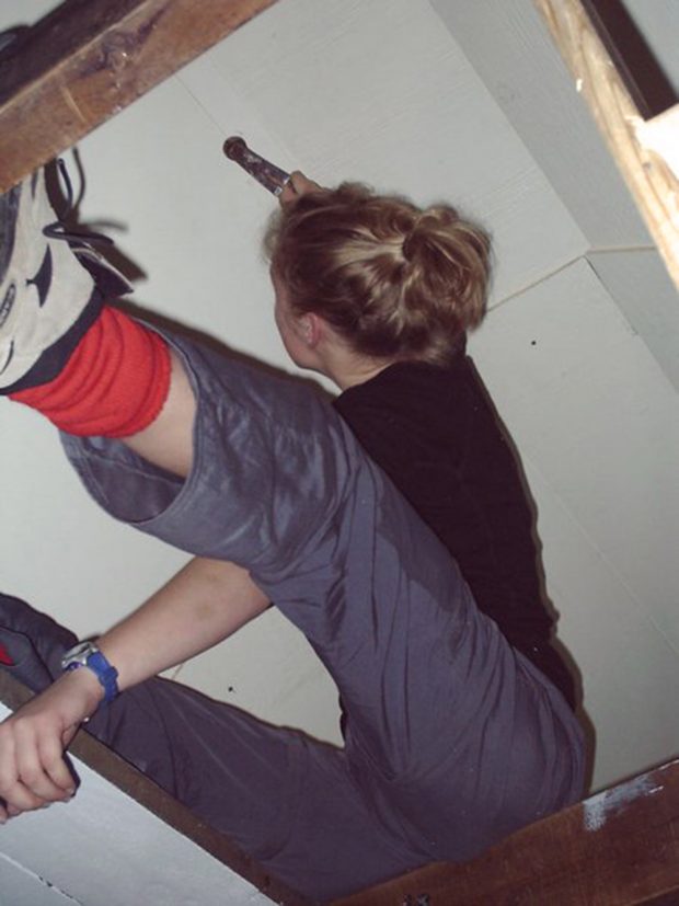 A woman wielding a hammer sits awkwardly on a roof support beam while reattaching a section of the interior wall of the Hut.