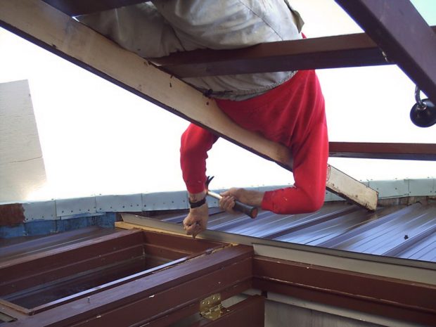 A man wearing a red shirt and light brow pants stands on a brown wooden ladder while attaching a piece of brown exterior aluminum siding.