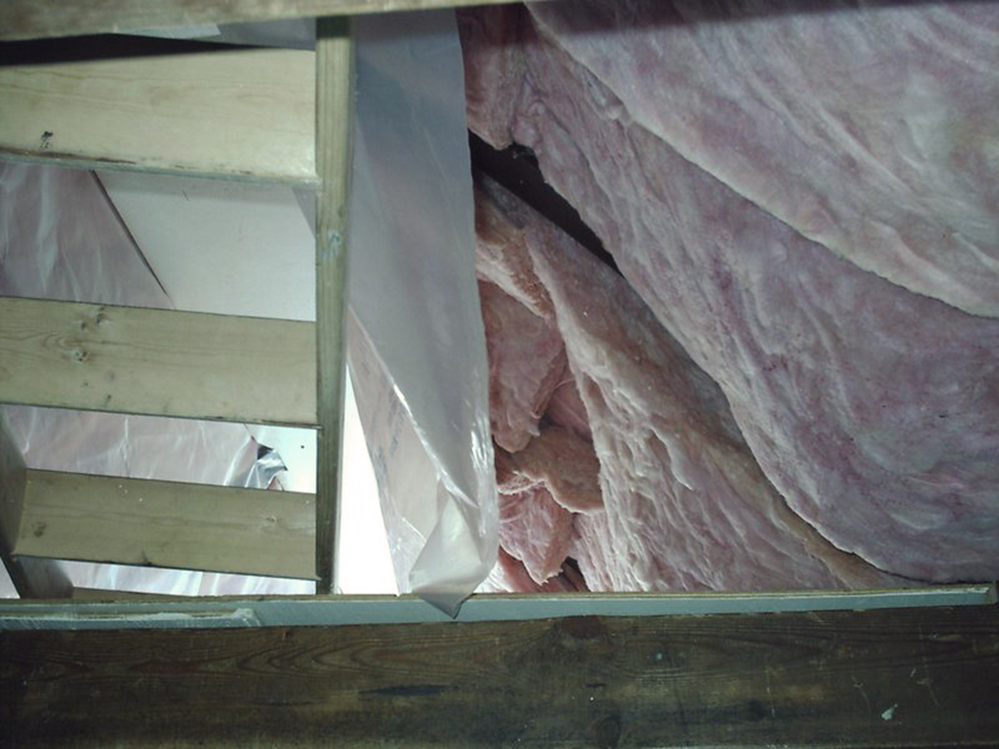 Un parement isolant est accroché au toit de la cabane et une feuille de plastique est fixée sur un côté, mais pas sur l’autre. Une échelle en bois vide est adossée contre une poutre en bois.