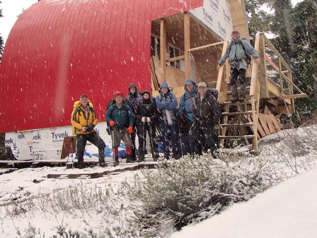 Group portrait of the construction crew that helped build the Hut with snow falling and lumber leaning against an end wall behind them on the large front porch.