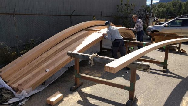 A man wearing a dark blue cap, light blue long-sleeved shirt and another man in a plaid shirt are moving one of the arched beams into place on its storage rack.