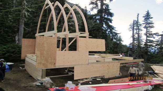 A side view of the arched frame being built with 4 complete arches in place and a worker busy joining a fifth arch at the far end of the structure.