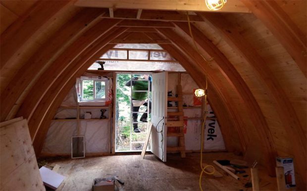 The second story loft space displays the laminated arches of the hut. Out the open second story door is a man standing on an aluminum ladder with evergreen trees in the background.