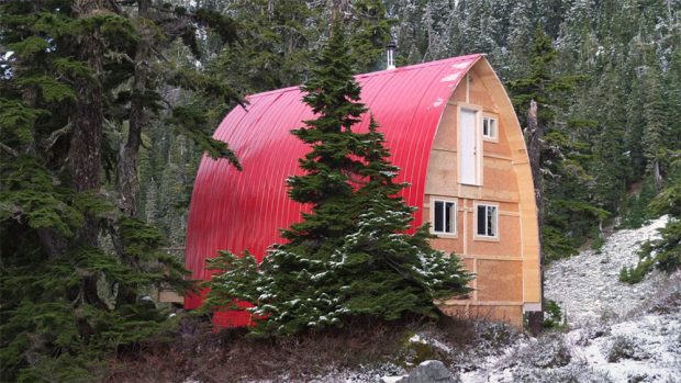 The bright red aluminum siding covering the roof of the arched Hut stands out against the dark green evergreen trees next to the hut. A light dusting of snow covers the ground and trees near the hut.