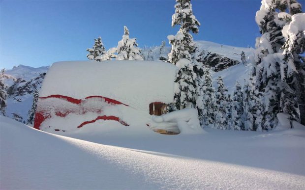 Deep snow surrounds the Hut and a portion of the red aluminum siding is uncovered. The winter sky is clear blue and snow covered evergreens are visible in the background.